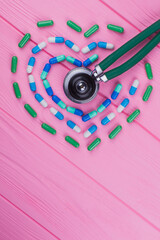 Capsules pills in a heart form and stethoscope. Pink wooden table surface. Topview flat lay.