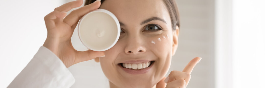 Close up wide image smiling young woman with smooth skin holding white plastic cream jar, head shot portrait beautiful girl looking at camera, enjoying skincare routine, applying moisturizing lotion