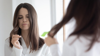 Mirror reflection unhappy young woman dissatisfied by dry hair with split ends, annoyed girl wearing white bathrobe standing in bathroom, touching messy hairs, worried about beauty problem