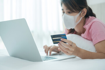 Sickness woman with face mask holding credit card and using computer laptop for online shopping on bed in morning at home. New normal, technology, ecommerce, digital banking and online payment concept