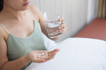 Adult woman holding pill and glass of water, taking medicine on bed in morning at home. Migraine, painkiller, headache, influenza, illness, sickness and healthcare concept