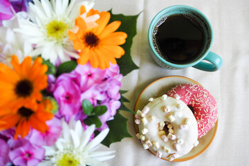 two donuts with pink icing and white with marshmallows on a brown round plate with coffee in a turquoise mug and a bouquet of flowers on a beige background top view