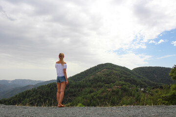 Naklejka premium A girl in shorts stands back and looks at the Troodos mountains from the observation platform of the Kikk monastery. Cyprus.