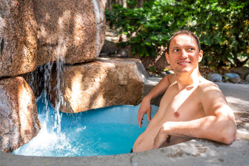 Young happy smiling man swimming in Japanese spa by stone pool colorful blue water and waterfall in Japan onsen