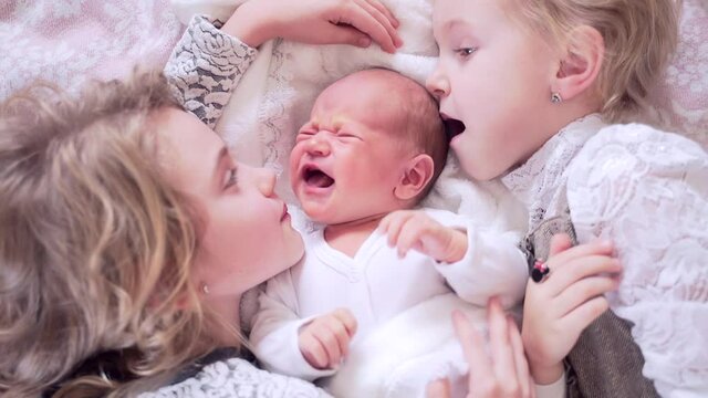 Three cute little children are lying on the bed. The sisters hug and kiss the newborn baby. Portrait of girls lying happy hugging close up. Happy and smiling together