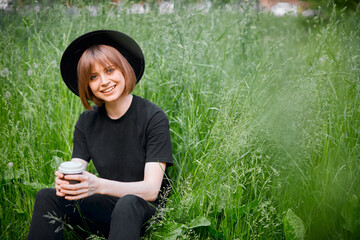 Cute young smiling girl in black and hat with a Cup of coffee in the green grass, eco-friendly active lifestyle