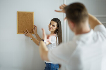 Woman helps her partner to hang painting on wall of their new house, she is framing the photo with her fingers