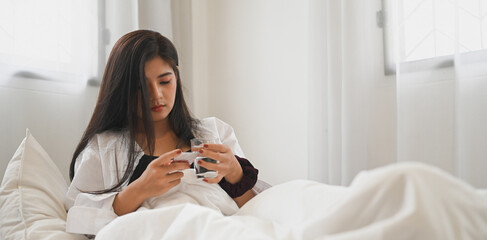 An ill woman is holding a glass of water after consuming a pill at the bed.