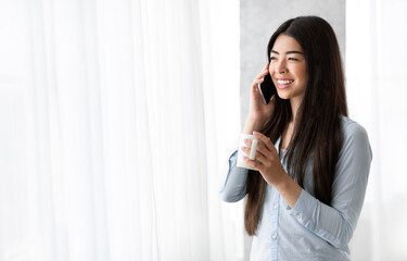 Pleasant call. Asian girl talking on cellphone and drinking coffee near window