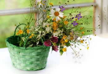 Fototapeta na wymiar beautiful bouquet of bright wild flowers in a green basket on the table against the window background