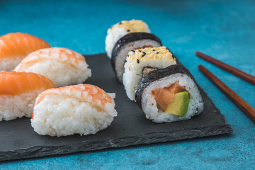 Variety of nigiri and maki shushi on slate and wooden chopsticks on blue background