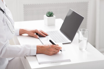 Online communication with patient and notes. Female doctor writes in notebook at table with laptop