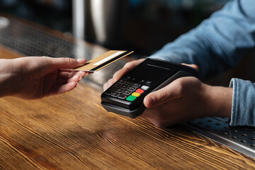 Modern contactless payment of bill in pub. Client hand holds credit card, bartender holding terminal - obrazy, fototapety, plakaty