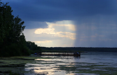 Storm clouds before the rain. Beautiful grey sky. Dramatic sky. A lone fisherman on the pier. Beyond the distant forest, it is raining, covering half the sky. Beautiful sunset