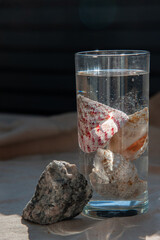 Sea shells inside a transparent glass with water on a sand-colored linen tablecloth. Nearby lies a gray granite stone. View from above. Close up