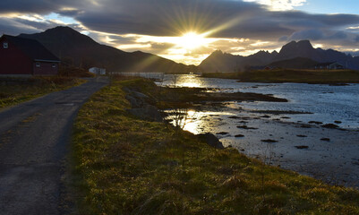 Obraz premium beautiful landscape of sunset,sunburst mountainranges,red norwegian house.The road have potholes.Sunlight reflect on the grass field.Fredvang lofoten norway.