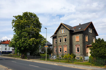 Fototapeta na wymiar Altes schiefernes Haus mit braunen Rolladen steht an der Hauptstraße umgeben von grünen Bäumen und Büschen. Bayern, Deutschland. 