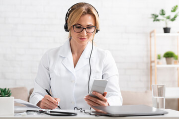 Writes patients to visit. Woman in white coat with headphones, smartphone and laptop makes notes in notebook
