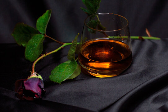 A glass of whiskey with a dark red rose on a black satin background
