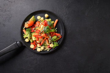 Frozen salad of vegetables in frying pan on black background. View from above. Copy space.
