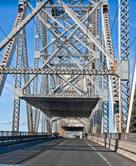 This vertical traffic image is a steel truss bridge from inside the bridge perspective.  Road has light traffic traveling on it in the daytime with bright blue skies.