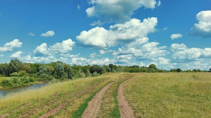country road in a field near the river against a beautiful blue sky with clouds