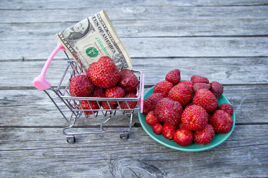 Red fresh strawberries in a trolley with a dallar banknote. Nearby stands a green saucer with strawberries on a wooden background.View from the side. Selling fruits