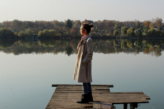 Young girl wearing a beige trench coat, jeans and black boots, standing on a wooden bridge and looking to the beautiful landscape. Young girl standing near the lake. People, nature, travel concept. - Powered by Adobe