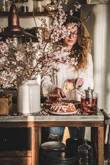 Spring or summer tea and cake setting. Young blond woman serving rose and almond gluten-free bundt cake with rose flowers on concrete kitchen counter with black tea and blooming branches in vase