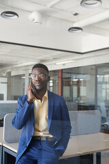 Vertical portrait of successful African-American businessman speaking by smartphone while leaning on desk in modern office interior, copy space