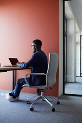Full length side view at contemporary African-American man using laptop at desk while working in...