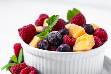 Frozen fruits: mango, raspberry, blueberry with fresh green mint in white bowl. Concept of healthy eating, delicious low calories dessert, summer refreshing light meal. White background copy space