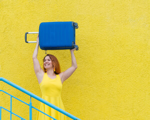 Happy woman in a yellow dress holds a blue suitcase over her head against the background of a...