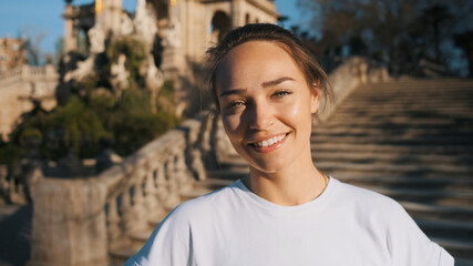 Portrait of beautiful smiling girl in white t-shirt happily looking in camera resting in city park with old architecture