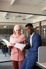 Vertical portrait of modern Muslim businesswoman talking to African-American colleague and...