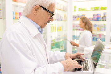 Senior male pharmacist searching for medicine in computer base by workplace