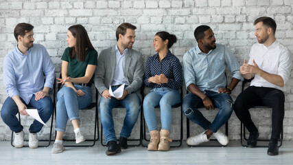 Group of multi ethnic people gathered in office corridor sitting on chairs in line talking with competitors while waiting turn job interview. HR, recruiting agency participants job-seekers concept - obrazy, fototapety, plakaty