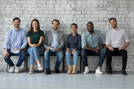 Six skilled potential company employees sit in queue in line smile looking at camera. Multi ethnic young people applicants wait for job interview feels confident. Human resources and staffing concept