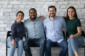 Four multi ethnic students friendly office employees sit together on chairs embracing smiling looking at camera. Warm relations between company members, amity and friendship, racial equality concept - obrazy, fototapety, plakaty