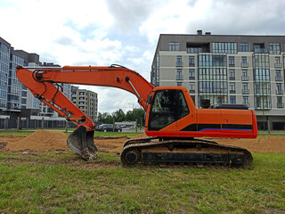 A yellow crawler excavator with a lowered bucket stands on the grass. Apartment buildings in the background.