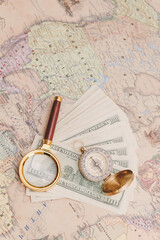 A bundle of American dollars lies with a compass and a magnifying glass on the world map