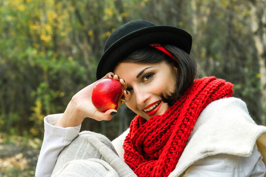 Positive woman in black hat and red scarf with a red apple in her hand smiles and look at the camera.