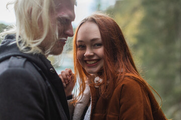 The happy couple have fun in forest. Close-up. Smiling, red-haired woman looks into the frame, a young man with blond hair looks at her