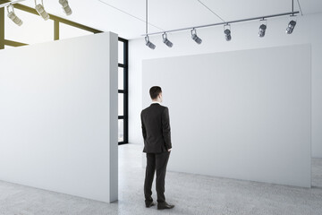 Businessman standing in clean gallery interior and looking on blank wall.