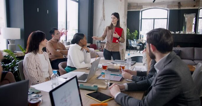 Young beautiful serious brunette female boss leading multiethnic office meeting seminar in loft modern light coworking.