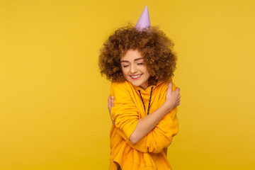 Portrait of selfish curly-haired hipster girl with funny cone hat embracing herself and smiling with pleasure expression, self-loving and complacency concept. studio shot isolated on yellow background