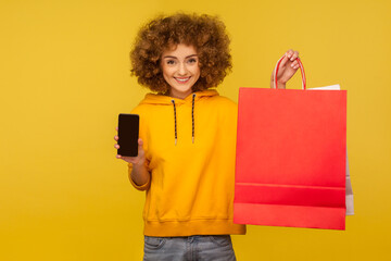 Portrait of optimistic curly-haired hipster girl in hoodie holding cellphone and shopping bags with blank area, copy space on packages for advertise. indoor studio shot, commercial of fashion store