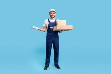 Full length happy delivery man in blue uniform with gloves holding cardboard boxes and showing copy space for advertise. Profession of courier, shipment and cargo transportation service. studio shot