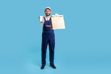 Full length smiling professional courier in blue uniform carrying groceries bag and showing mobile phone with mock up display for advertise image. App of food delivery service. studio shot isolated
