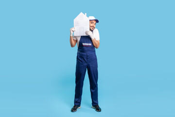 Full length cheerful handyman, building worker in uniform peeking out of paper house and smiling to camera. Mover guy offering help in relocation, home repair services. indoor studio shot isolated